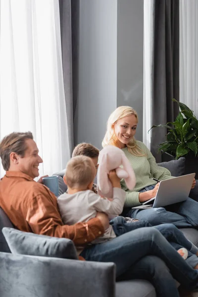 Lächelnde Frau arbeitet auf der Couch am Laptop, während ihr Mann mit Söhnen spielt — Stockfoto