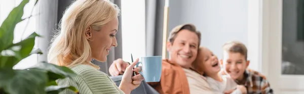 Glückliche Frau trinkt Tee in der Nähe verschwommener Familie im Wohnzimmer, Banner — Stockfoto