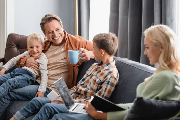 Hombre feliz con taza de té sentado en el sofá con los hijos, mientras que la esposa borrosa trabajando en el ordenador portátil - foto de stock