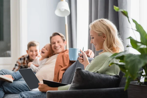 Lächelnde Frau mit einer Tasse Tee, die mit Laptop und Notizbuch in der Nähe einer verschwommenen Familie auf dem Sofa arbeitet — Stockfoto