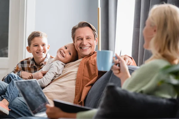 Cheerful man with sons near blurred woman with cup of tea and laptop on sofa — Stock Photo