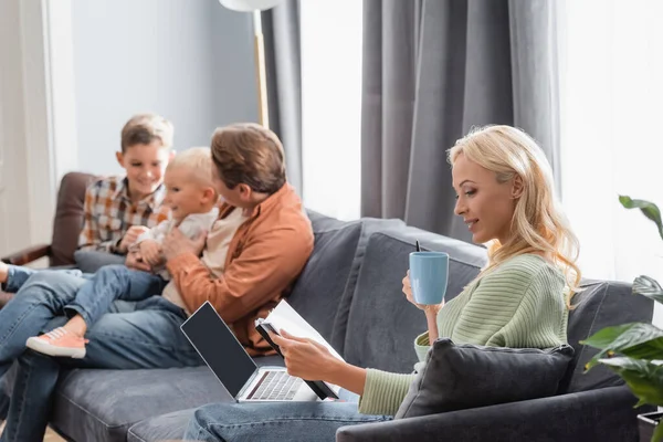 Frau mit Notizbuch und Laptop arbeitet auf Sofa neben verschwommenem Ehemann mit Söhnen — Stockfoto