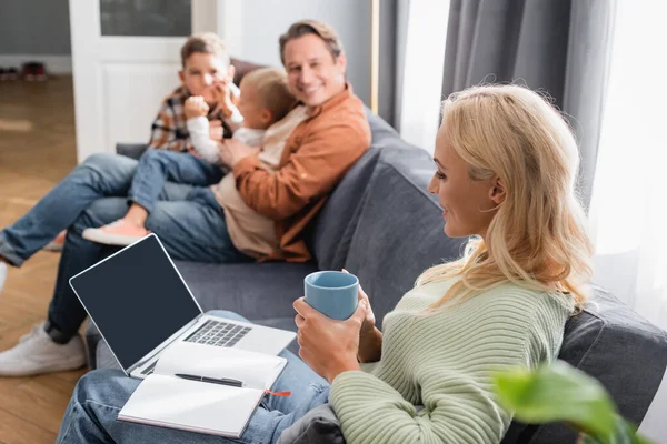 Femme avec ordinateur portable, ordinateur portable et tasse de thé assis sur le canapé près de famille joyeuse floue — Photo de stock