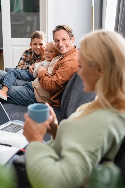 Femme floue avec tasse à thé travaillant sur ordinateur portable près mari heureux avec des fils sur le canapé — Photo de stock