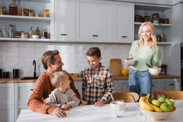 Mujer sonriente sosteniendo cuencos cerca de marido e hijos sentados en la mesa de la cocina - foto de stock