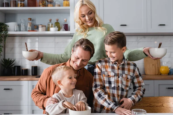 Lächelnde Frau mit Schalen neben Mann und Söhnen, die auf das Frühstück warten — Stockfoto