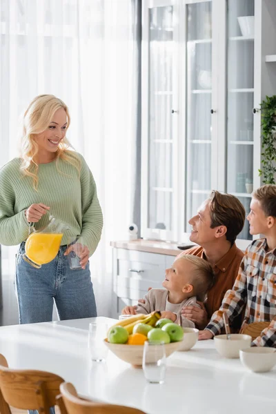 Cheerful woman holding orange juice near family having breakfast in kitchen — Stock Photo