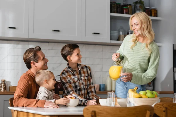 Femme gaie verser du jus d'orange pendant le petit déjeuner avec le mari et les enfants — Photo de stock