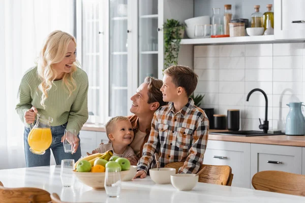 Mujer alegre sosteniendo jugo de naranja cerca de marido e hijos durante el desayuno — Stock Photo