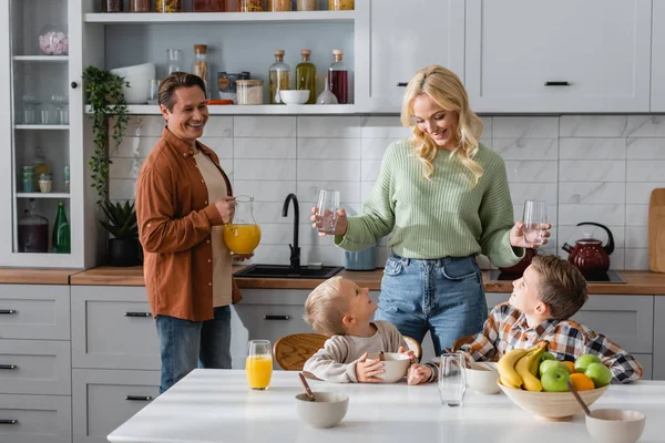 Feliz pareja sosteniendo vasos y jarra con jugo de naranja cerca de los niños desayunando en la cocina - foto de stock