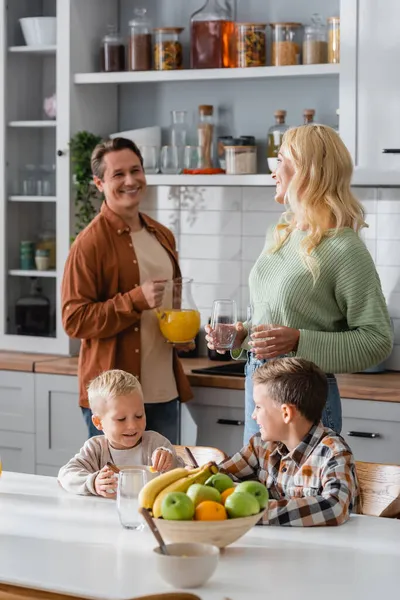 Hermanos desayunando cerca de padres borrosos con vasos y jugo de naranja — Stock Photo