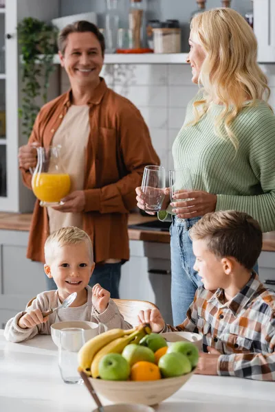 Lächelnde Eltern schauen einander an, während sie Gläser und Orangensaft in der Nähe von Söhnen beim Frühstück halten — Stockfoto