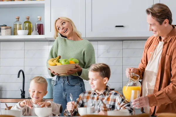 Mulher com tigela de frutas frescas falando no smartphone perto do homem com suco de laranja e meninos tomando café da manhã — Fotografia de Stock