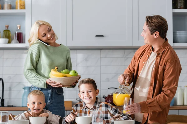 Lächelnde Frau mit frischem Obst spricht auf Smartphone neben Ehemann, der beim Frühstück Orangensaft einschenkt — Stockfoto