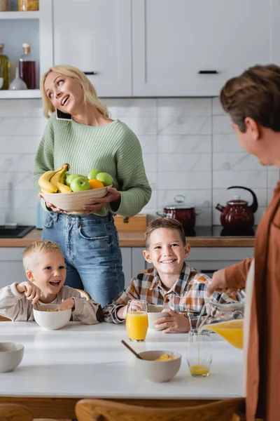 Femme avec des fruits parlant sur téléphone portable et homme versant du jus d'orange près des garçons prenant le petit déjeuner dans la cuisine — Photo de stock