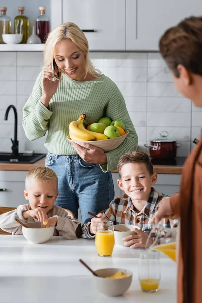 Mujer con frutas frescas hablando en el teléfono móvil cerca del marido borroso y los niños desayunando — Stock Photo