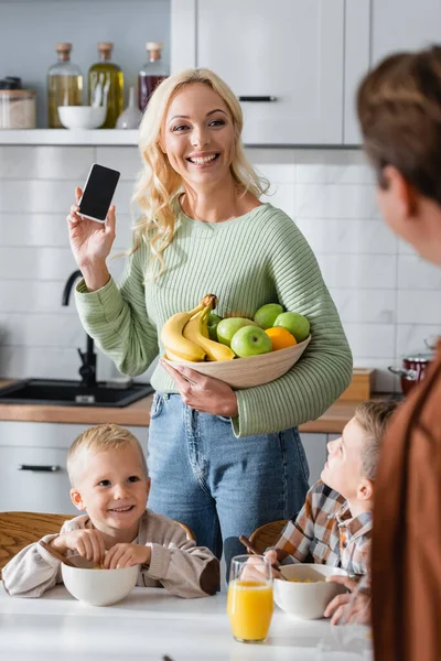 Happy woman holding smartphone with blank screen and fresh fruits near kids and blurred husband in kitchen — Stock Photo