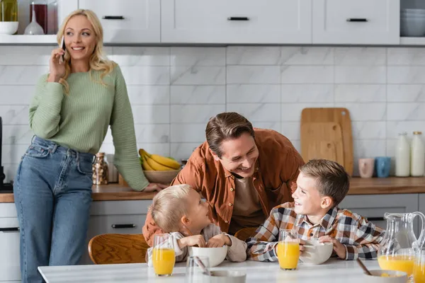 Hombre alegre hablando con los hijos desayunando cerca de la esposa con el teléfono móvil sobre fondo borroso - foto de stock