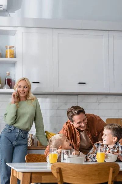 Femme floue et heureuse parlant sur téléphone mobile près du mari avec des fils prenant le petit déjeuner dans la cuisine — Photo de stock