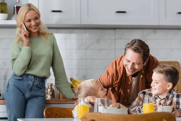 Mujer borrosa hablando por teléfono móvil cerca de marido feliz de pie cerca de los hijos desayunando en la cocina - foto de stock