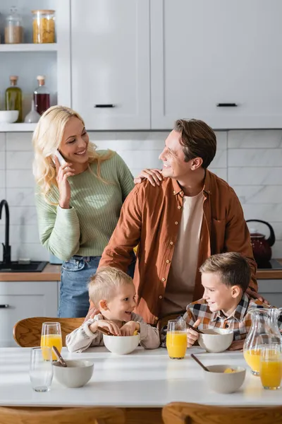 Mujer sonriente hablando por teléfono móvil mientras busca marido y niños durante el desayuno - foto de stock