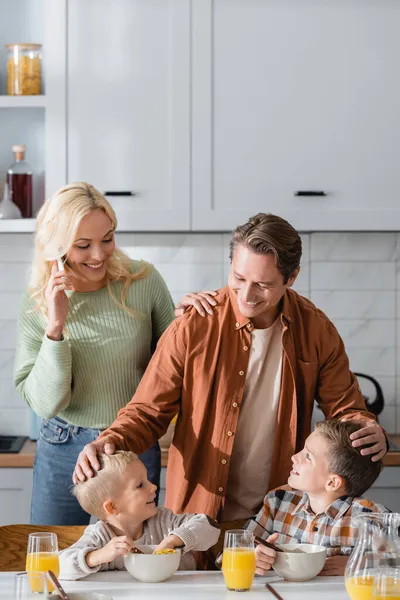 Glücklicher Vater berührt Köpfe von Söhnen beim Frühstück in der Nähe von Mama, die mit dem Handy telefoniert — Stockfoto