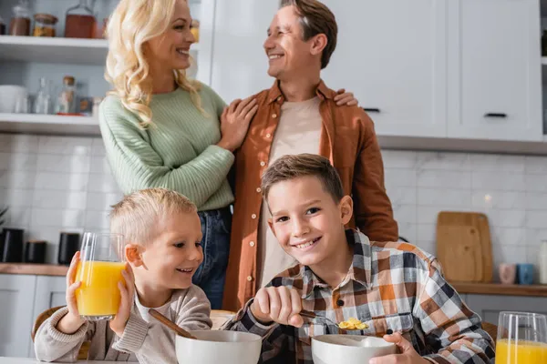 Fröhliche Brüder beim Frühstück in der Nähe von Eltern, die sich auf verschwommenem Hintergrund umarmen — Stockfoto