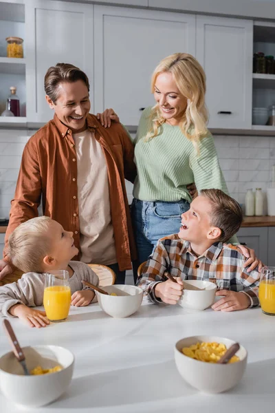 Счастливая пара рядом сыновья едят хлопья кукурузы и пить апельсиновый сок во время завтрака — стоковое фото