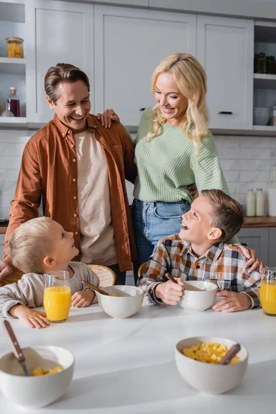 Couple heureux près de fils manger des flocons de maïs et boire du jus d'orange pendant le petit déjeuner — Photo de stock