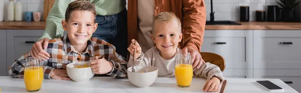 Счастливые мальчики едят вкусные хлопья кукурузы возле апельсинового сока и родителей, баннер — стоковое фото