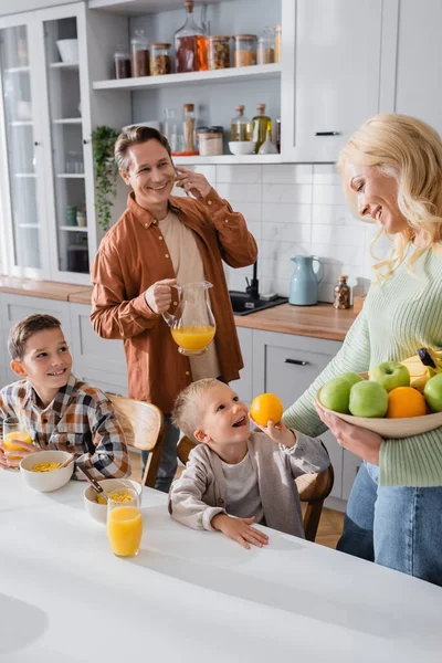 Smiling man with orange juice talking on mobile phone near family in kitchen — Stock Photo