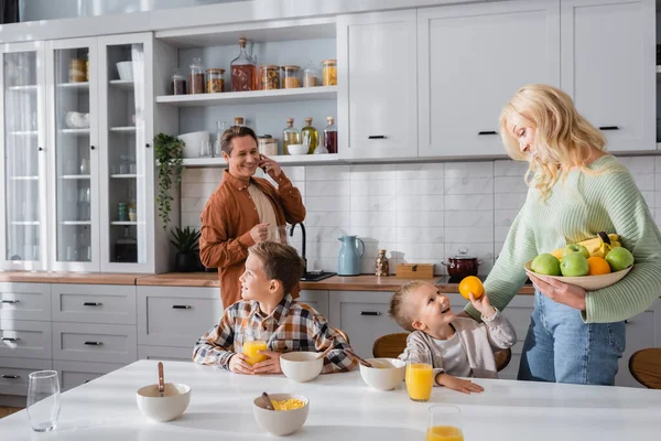 Uomo sorridente che parla su smartphone mentre moglie tiene frutta vicino ai bambini in cucina — Foto stock