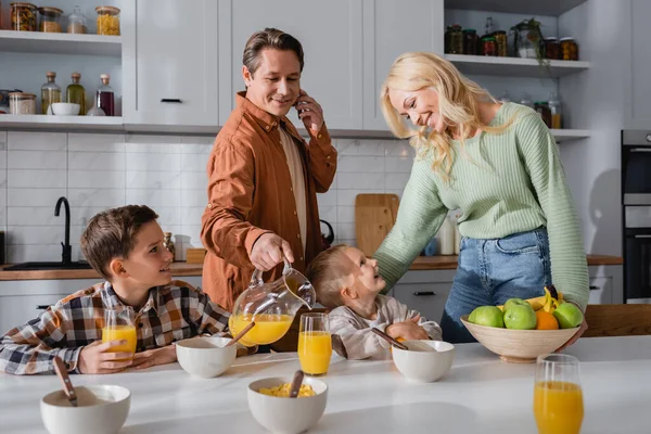 Mann spricht mit Smartphone, während er beim Frühstück mit Familie Orangensaft einschenkt — Stockfoto