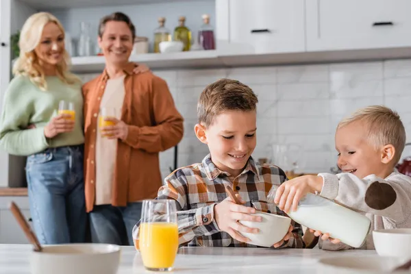 Garçon verser du lait dans le bol tout en prenant le petit déjeuner avec le frère près des parents flous — Photo de stock