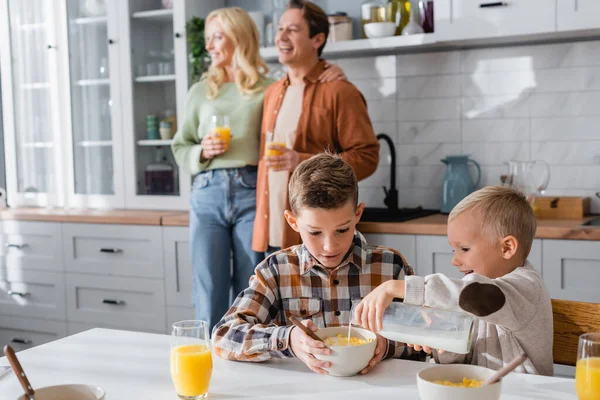 Niño vertiendo leche en copos de maíz mientras desayunaba con su hermano cerca de la familia borrosa - foto de stock