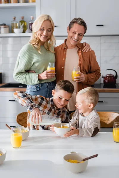 Heureux couple regardant fils verser du lait pendant le petit déjeuner avec frère — Photo de stock