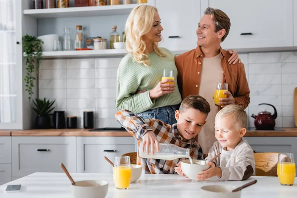 Coppia allegra con occhiali di succo d'arancia vicino a bambini che fanno colazione in cucina — Foto stock