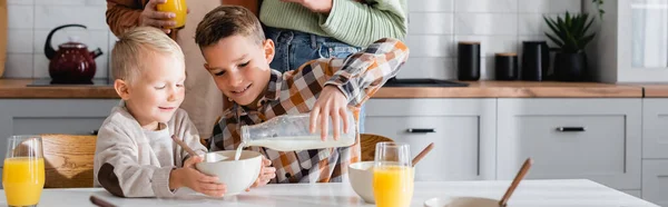 Niño vertiendo leche durante el desayuno con el hermano cerca de los padres, pancarta - foto de stock