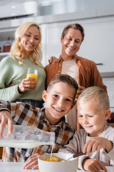Smiling kid looking at camera while pouring milk into bowl with delicious corn flakes — Stock Photo