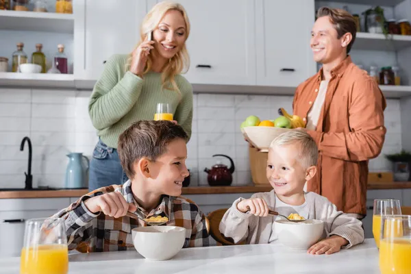 Femme floue parlant sur téléphone mobile près du mari tenant des fruits et des enfants prenant le petit déjeuner — Photo de stock