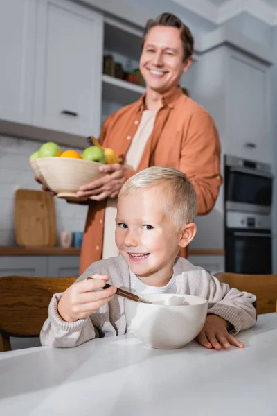 Menino sorrindo olhando para a câmera enquanto toma café da manhã perto de pai desfocado com frutas frescas — Fotografia de Stock