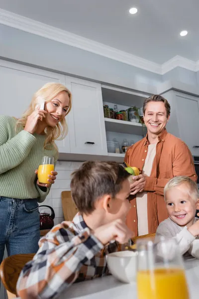 Frau hält Glas Orangensaft in der Hand und telefoniert mit Smartphone in der Nähe der Familie beim Frühstück in der Küche — Stockfoto