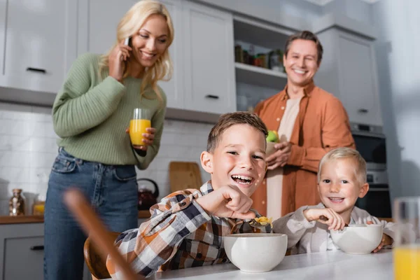 Allegri ragazzi che fanno colazione vicino sfocato padre e mamma parlando su smartphone — Foto stock