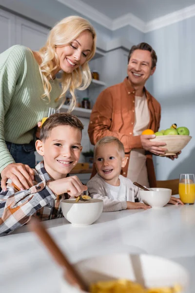 Irmãos felizes comendo flocos de milho perto de pais felizes na cozinha — Fotografia de Stock