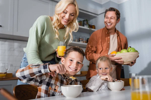 Frères joyeux manger des céréales pour le petit déjeuner près des parents avec des fruits frais et du jus d'orange — Photo de stock