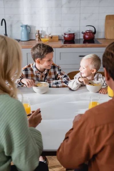 Lächelnde Jungen frühstücken, während sie Cornflakes in der Nähe verschwommener Eltern essen — Stockfoto