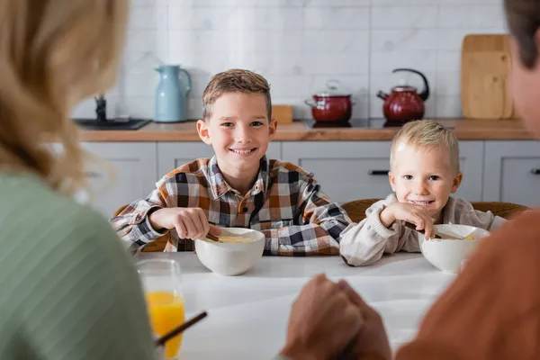 Lächelnde Jungen essen leckere Cornflakes in der Nähe verschwommener Eltern in der Küche — Stockfoto