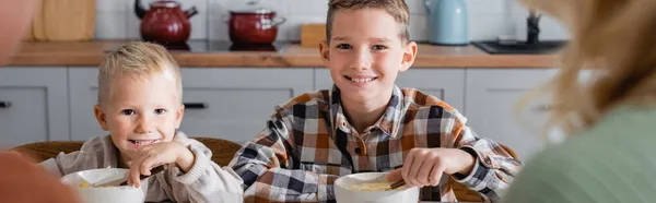 Frères gais manger des flocons de maïs savoureux pour le petit déjeuner près des parents flous, bannière — Photo de stock