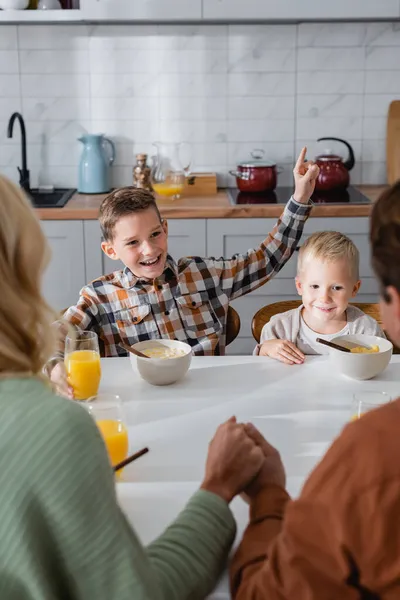Happy boy pointing with finger during breakfast with brother and parents holding hands on blurred foreground — Stock Photo