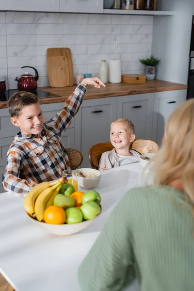 Sorrindo menino apontando com o dedo para o irmão durante o café da manhã com cereais e frutas frescas ner mãe turva — Fotografia de Stock